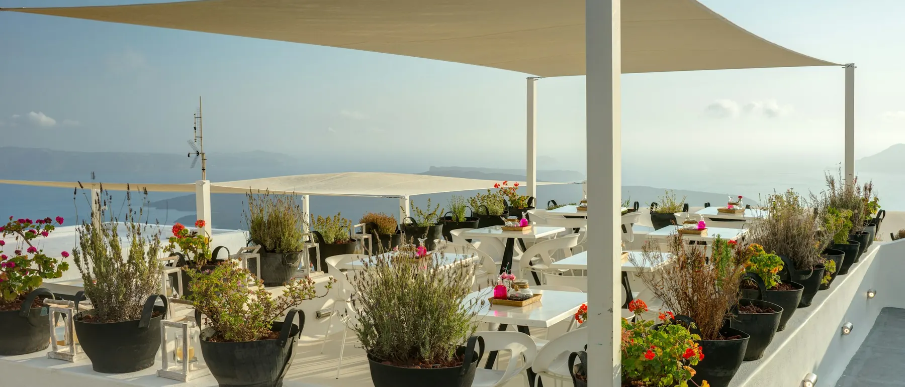 An empty terrace restaurant with white tables, a sail canopy and potted herbs, looking out over the sea