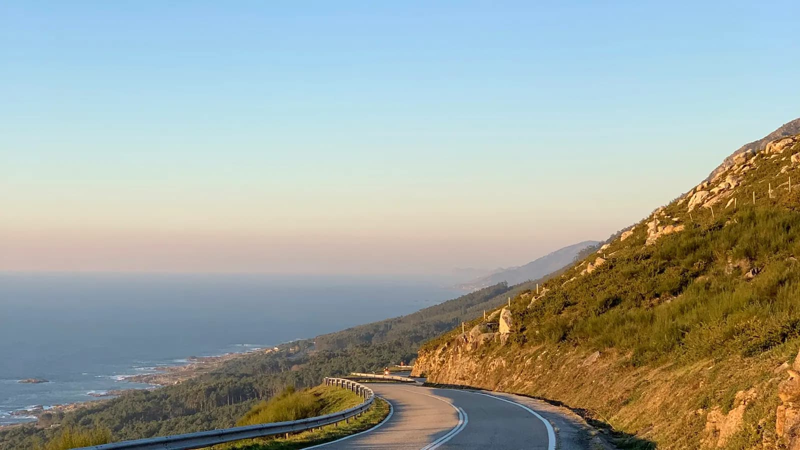 An empty road winding along a hillside above the sea at sunset