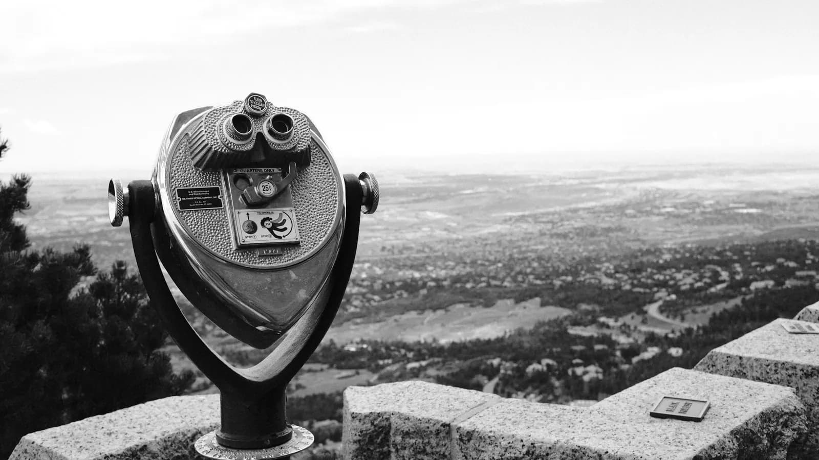 Coin-operated binoculars on a stone wall looking out over a wide valley, in black and white